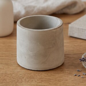 Könnte beinhalten: Ein zylindrischer Betonübertopf mit glatter, hellgrauer Oberfläche und hohlem Innenraum. Das Gefäß verfügt über einen flachen Boden und gerade Wände und bietet ein neutrales, industrielles Erscheinungsbild, das sich für kleine Pflanzen eignet.