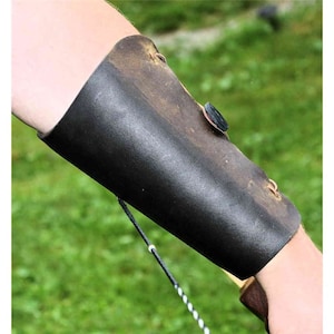 Può includere: Un bracciale da tiro con l'arco in pelle nera con chiusura a bottone e dettagli di cucitura. Il bracciale è indossato sul braccio sinistro e serve a proteggere il braccio dalla corda dell'arco.