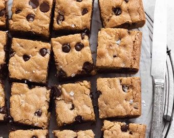 May include: A close-up of a baking sheet filled with squares of chocolate chip cookie bars. The bars are golden brown and have a light dusting of sea salt on top.