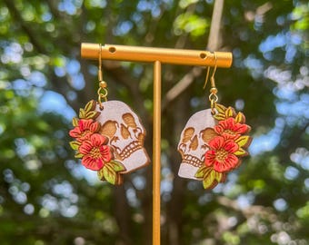 Puede incluir: Un par de pendientes con forma de calavera con ganchos dorados. Cada pendiente presenta un diseño de calavera con flores rojas y amarillas y hojas verdes. Los pendientes son de madera y cuelgan de un soporte dorado.