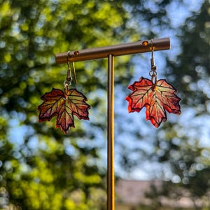 May include: A pair of leaf-shaped earrings. The earrings are designed to resemble autumn maple leaves, with red, orange, and yellow hues. The earrings hang from gold-colored hooks, and are displayed on a gold-colored stand against a blurred green background.