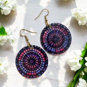 Può includere: Un paio di orecchini rotondi con un design a vetri colorati. Gli orecchini sono viola, rosa e blu con uno sfondo nero. Gli orecchini sono appesi a ganci dorati.