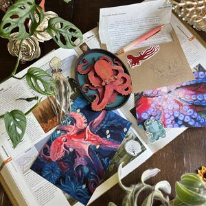 May include: A collection of octopus-themed items including a wooden octopus plaque, a pink pen, a brown envelope, a red octopus sticker, a teal octopus sticker, a tree sticker, and two octopus art prints resting on an open book with text.