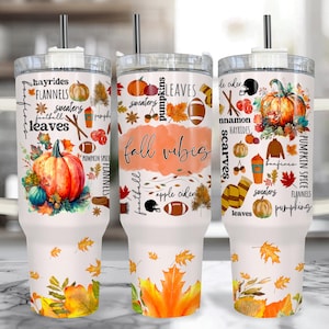 Może przedstawiać: Trzy białe kubki termiczne z uchwytami, ozdobione grafiką w jesiennym klimacie. Każdy kubek przedstawia dynie, liście oraz napisy "Fall Vibes", "Hayrides" i "Flannels". Wzory obejmują piłki nożne, swetry i jesienne kolory.