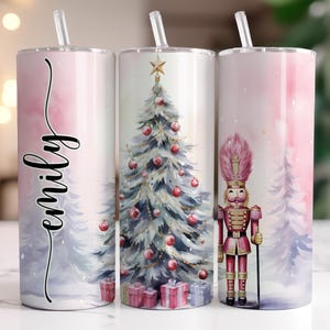 May include: Three stainless steel tumblers with straws. One tumbler features the name "emily" in black script. The other two tumblers have a Christmas tree and a nutcracker design. The tumblers have a pink and blue watercolor background.