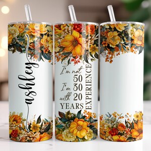 May include: A white tumbler with a floral design featuring yellow, orange, and green flowers. The tumbler has a straw and the name "Ashley" in cursive. The text "I'm not 50, I'm 30 with 20 years experience" is also printed on the tumbler.