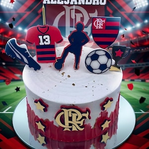 K&ouml;nnte beinhalten: Ein dekorierter Kuchen mit Fu&szlig;ball-Thema. Der Kuchen ist wei&szlig; mit roten und goldenen Akzenten und mit einem Fu&szlig;ball, einer Troph&auml;e, einem Trikot, einem Fu&szlig;ballschuh und einem Schild verziert. Der Name "ALEJANDRO" steht oben.