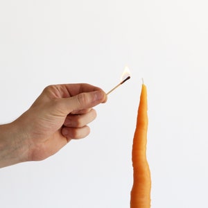 Puede incluir: Una mano sostiene una cerilla encendida cerca de una vela larga de color naranja con forma de zanahoria.