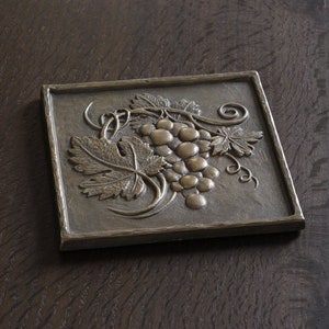 May include: A square, bronze-colored wall plaque with a raised design of a grapevine with leaves and grapes.