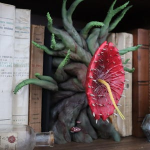 Op de afbeelding: Een handgemaakte, grillige sculptuur van een boom met een grote, rode, stekelige bloem. De boom heeft groene, tentakelachtige takken. De sculptuur staat op een plank met antieke boeken en een glazen fles.