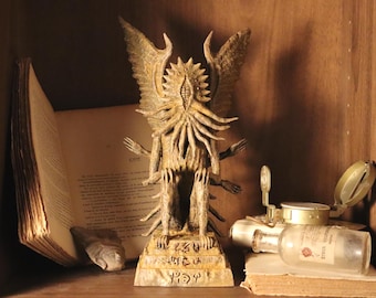 May include: A detailed, weathered statue of a winged creature with multiple arms, standing on a pedestal with engraved symbols. Beside it are an open antique book, a compass, and a small glass bottle, all arranged on a wooden shelf.
