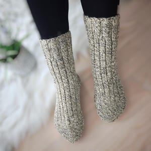 Könnte beinhalten: Ein Paar dicke, gerippte Stricksocken in Grau- und Cremetönen. Die Socken werden mit schwarzen Leggings getragen. Die Socken sind von den Knöcheln abwärts sichtbar.