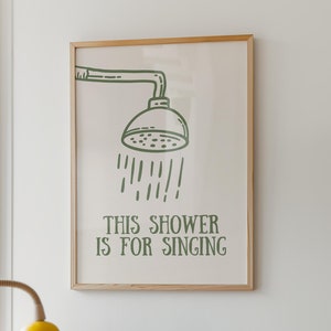 Op de afbeelding: Een ingelijst print met een groene illustratie van een douchekop en de tekst "This shower is for singing". De print hangt aan een witte muur boven een houten kast met een gele lamp, een stereo-installatie en een toetsenbord.