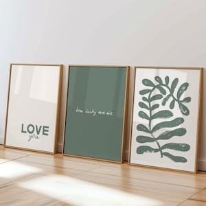 以下が含まれることがあります： 白い壁に立てかけられた3つの額入りアートプリント。最初のプリントには「LOVE you」の文字。2つ目はセージグリーンで「how lucky are we」と書かれています。3つ目は緑色の植物のデザインです。インテリアに最適です。