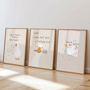 Puede incluir: Tres impresiones artísticas enmarcadas con ilustraciones de gansos de dibujos animados sobre un fondo beige claro. Las impresiones presentan el texto: "NO OLVIDES LAVARTE LAS MANOS", "¡MIRA... INCLUSO TE CONSEGUÍ UN CEPILLO DE DIENTES!" y "time to BATH!". Los marcos son de color marrón claro.