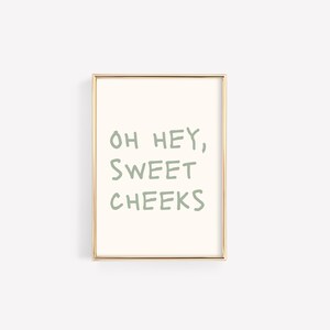Może przedstawiać: Wydruk w ramie ze złotą ramką i białym tłem. Tekst "OH HEY, SWEET CHEEKS" jest napisany na zielono.