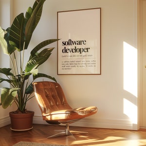 May include: A framed print with a white background and black text that reads "software developer" and a definition of the term. The print is hanging on a white wall in a room with a brown leather chair and a green plant in a brown pot.