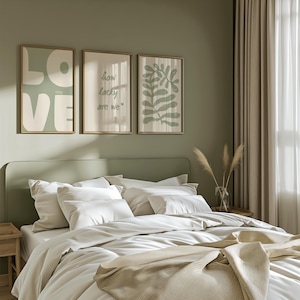 May include: Three framed prints on a sage green wall above a bed with white bedding. The prints feature a white graphic of the word "LOVE" with the word "YOU" in smaller letters, a green graphic with the text "how lucky are we", and a green abstract leaf design.
