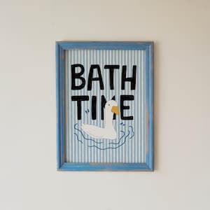 May include: A framed wall art print featuring a white goose with an orange beak swimming in blue water ripples. The background has vertical light blue and white stripes. The black text BATH TIME is printed in a bold, hand-drawn style font.