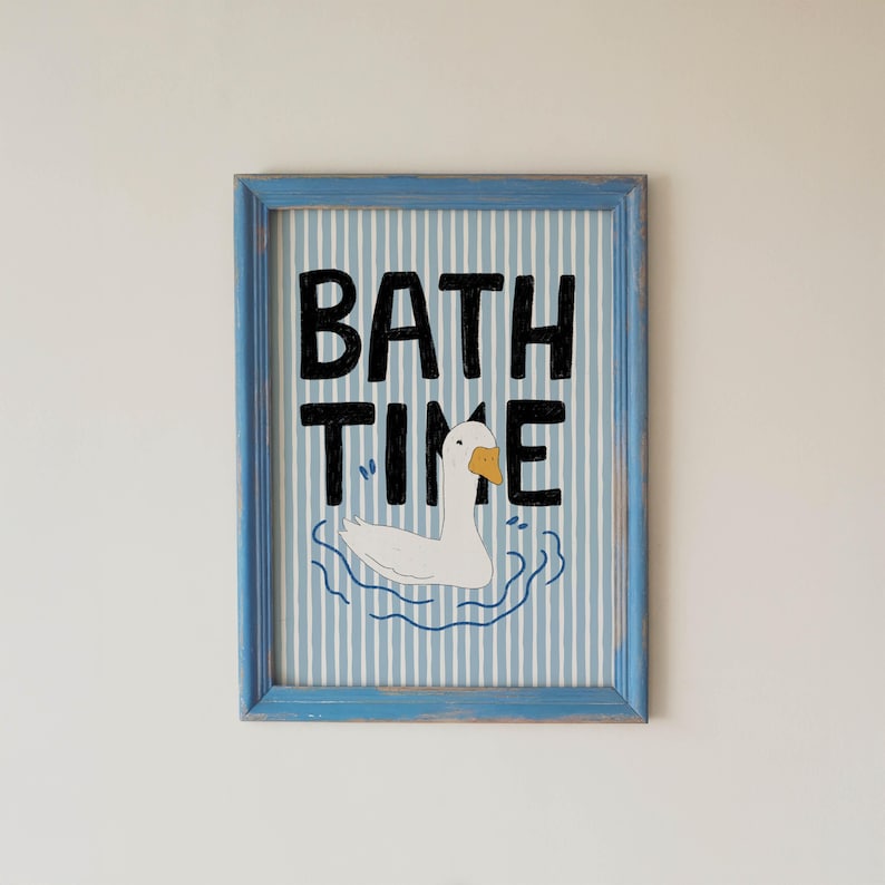 May include: A framed wall art print featuring a white goose with an orange beak swimming in blue water ripples. The background has vertical light blue and white stripes. The black text BATH TIME is printed in a bold, hand-drawn style font.