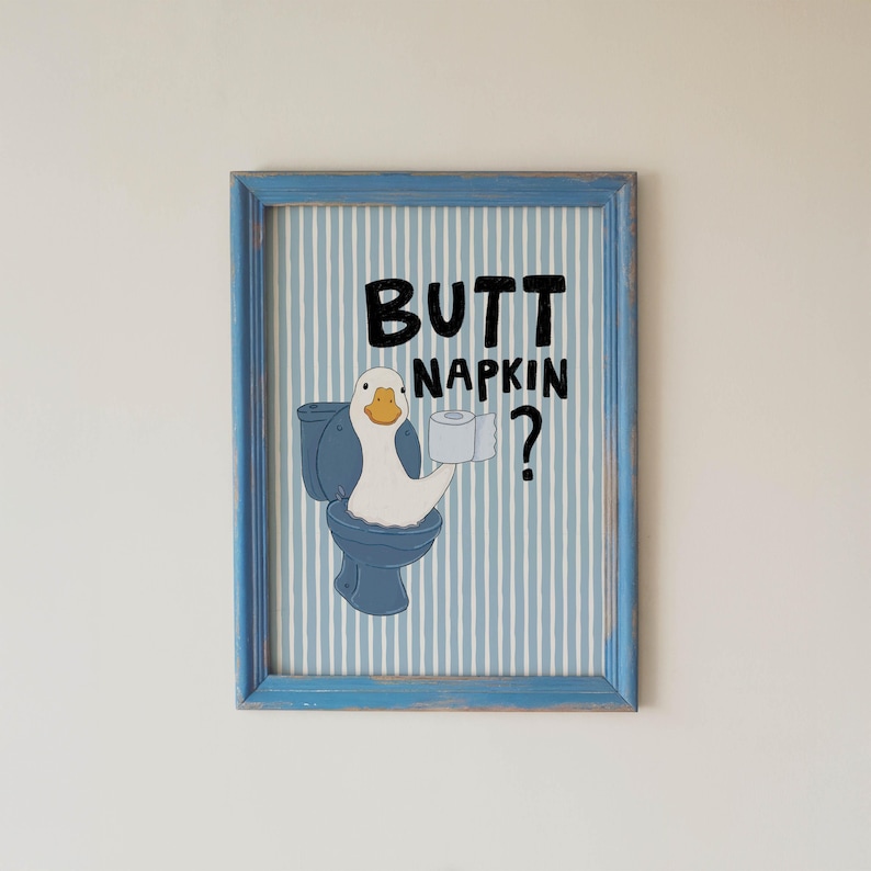 May include: A framed wall art print featuring a white goose sitting on a blue toilet, holding a roll of toilet paper. The background has vertical white and blue stripes. The black text reads BUTT NAPKIN? The frame is light blue with a distressed finish.