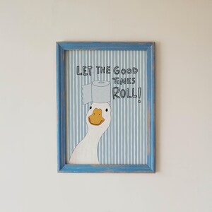 May include: A framed wall art print featuring a white goose with an orange beak balancing a roll of toilet paper on its head. The background has vertical light blue and white stripes. The text reads: LET THE GOOD TIMES ROLL!