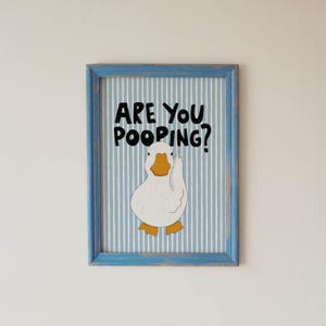 May include: A framed wall art print featuring a white duck with an orange beak and feet centered on a light blue and white vertical striped background. The black text above the duck reads ARE YOU POOPING? The frame is a distressed light blue wood.