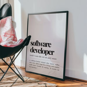 May include: A black framed print with a white background and black text that reads "software developer" and a definition of the term. The print is hanging on a white wall above a black chair with a pink and white blanket.