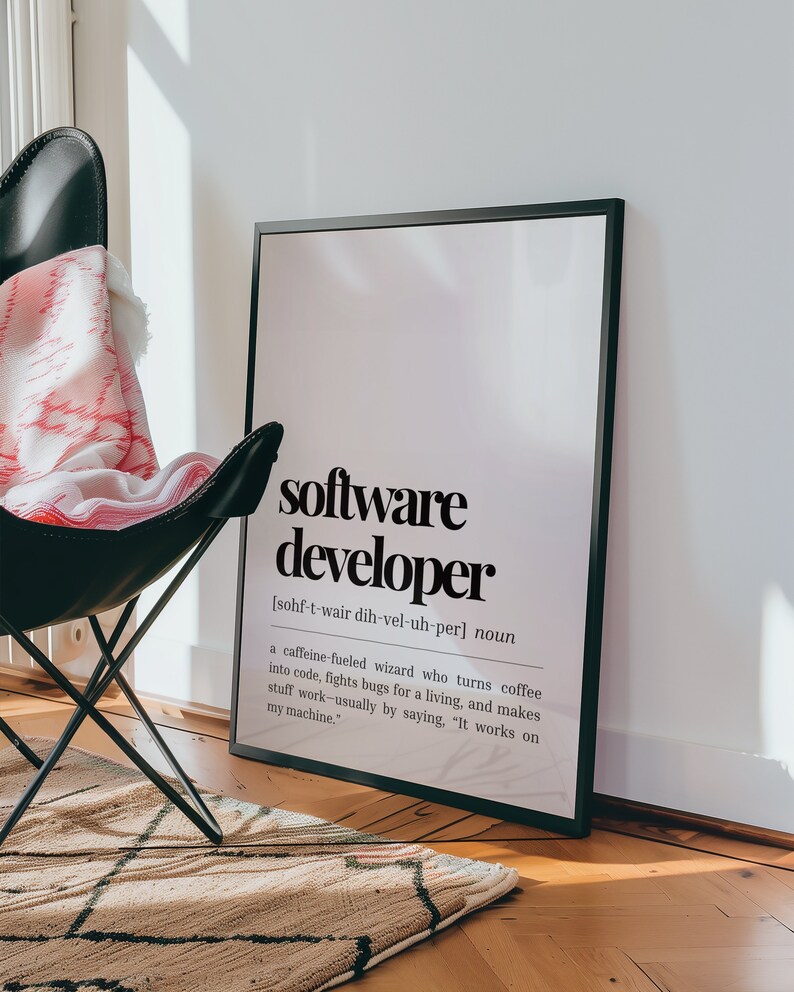 May include: A black framed print with a white background and black text that reads "software developer" and a definition of the term. The print is hanging on a white wall above a black chair with a pink and white blanket.