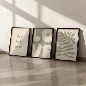 May include: Three framed prints with a minimalist design. The first print has the text "how lucky are we." The second print has the word "LOVE" in large, green letters with the word "YOU" in smaller letters below. The third print has a green abstract leaf design.