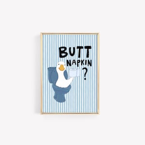 May include: A framed wall art print featuring a white goose sitting on a blue toilet, holding a roll of toilet paper. The background has light blue and white vertical stripes. The text reads BUTT NAPKIN? in bold black letters above the goose.