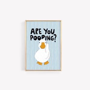 May include: A rectangular wall art print in a gold frame featuring a white duck with an orange beak and feet against a light blue and white vertical striped background. The black text above the duck reads, ARE YOU POOPING? in a bold, hand-drawn style.