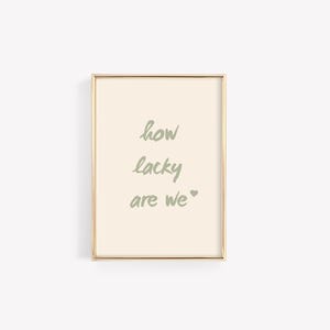 May include: A framed print with a light beige background and gold frame. The text "how lucky are we" is written in green.