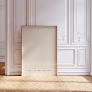 May include: A gold framed poster with a white background and the text "YOU ARE MY SUNSHINE" in red. The poster is leaning against a white wall with ornate molding.