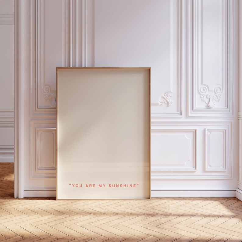 May include: A gold framed poster with a white background and the text "YOU ARE MY SUNSHINE" in red. The poster is leaning against a white wall with ornate molding.