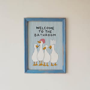 May include: A framed wall print featuring four white ducks on a light blue and white vertical striped background. The text WELCOME TO THE BATHROOM is printed in black. One duck wears a pink shower cap, one holds a toothbrush, and one has toilet paper.