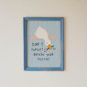 May include: A framed wall art print featuring a white duck with an orange beak holding a blue toothbrush with red toothpaste. The background has vertical light blue and white stripes. The text reads: DON'T FORGET TO BRUSH YOUR TEETH!
