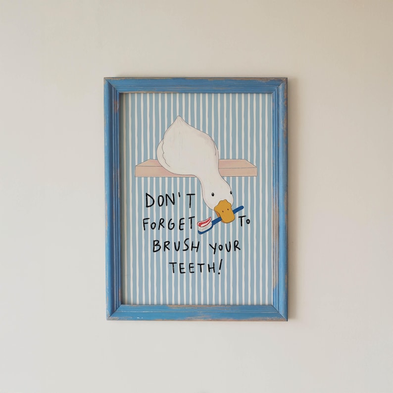 May include: A framed wall art print featuring a white duck with an orange beak holding a blue toothbrush with red toothpaste. The background has vertical light blue and white stripes. The text reads: DON'T FORGET TO BRUSH YOUR TEETH!