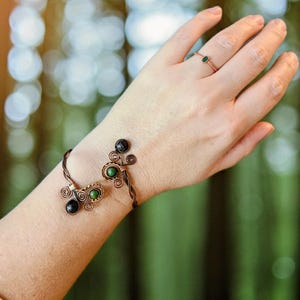 May include: A copper wire bracelet with black and green gemstone accents. The bracelet features a swirling design with a matching ring with a green gemstone. The jewelry is displayed on a person's arm.