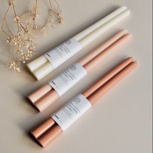 Może przedstawiać: Trzy pary świec taper w kolorze białym, jasnor&oacute;żowym i brązowym. Świece są zapakowane w biały papier z tekstem "Pair of Classic Taper Dining Candles" i nazwą marki "AJORA".