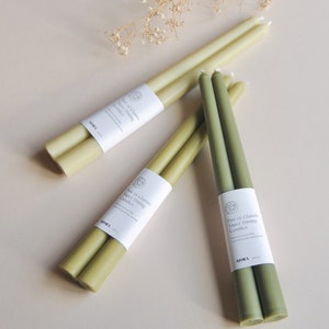 Może przedstawiać: Zestaw trzech par zielonych i oliwkowych świec stożkowych. Każda para jest zapakowana w biały papier z tekstem "Pair of Classic Taper Dining Candles" i nazwą marki "MORA".