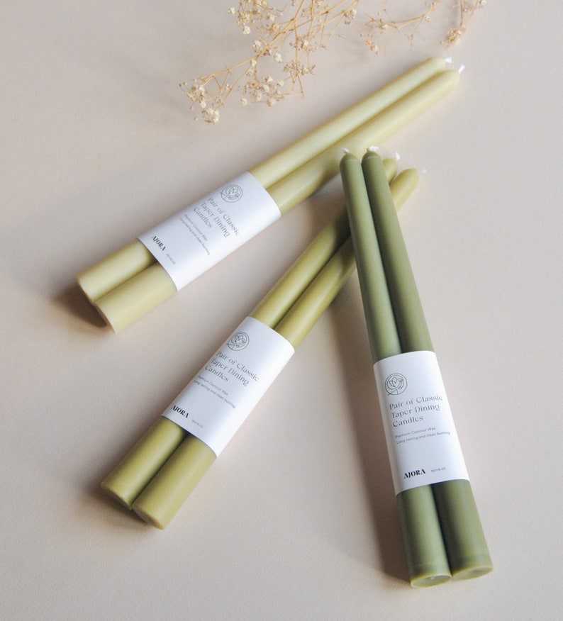 Może przedstawiać: Zestaw trzech par zielonych i oliwkowych świec stożkowych. Każda para jest zapakowana w biały papier z tekstem "Pair of Classic Taper Dining Candles" i nazwą marki "MORA".