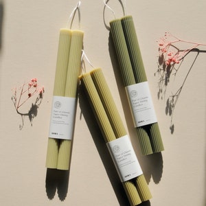 Może przedstawiać: Zestaw trzech par zielonych i beżowych świec taper. Każda para jest owinięta w papier z tekstem "Pair of Classic Taper Dining Candles" i słowem "MORA".