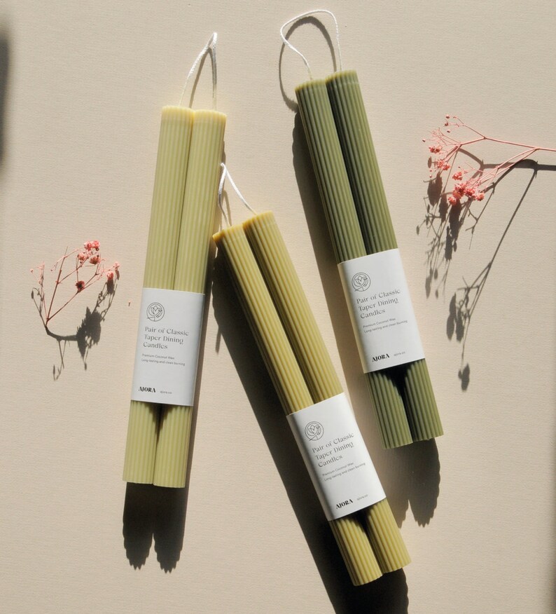 Może przedstawiać: Zestaw trzech par zielonych i beżowych świec taper. Każda para jest owinięta w papier z tekstem "Pair of Classic Taper Dining Candles" i słowem "MORA".