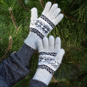 May include: A pair of light gray knit gloves with a black and white geometric pattern on the cuffs and back of the hands. The gloves are set against a backdrop of green pine needles.