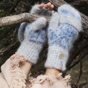 May include: Gray knitted mittens with blue snowflake patterns. The mittens have a button closure and a label that says "Siberia". The mittens are shown on a person holding a tree branch.