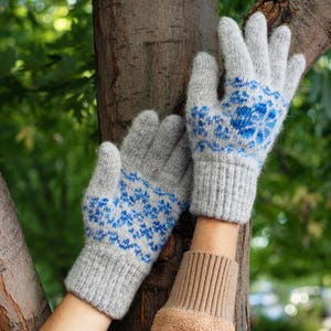 May include: A pair of gray knitted gloves with a blue floral pattern. The gloves are made of a soft, warm material and have ribbed cuffs. The gloves are displayed against a tree trunk with a green background.