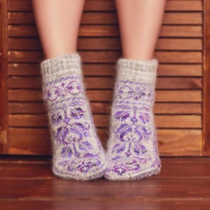May include: A pair of knitted socks with a floral pattern in shades of purple and white. The socks are worn by a person and are set against a wooden background. The socks have a gray cuff and a detailed design.