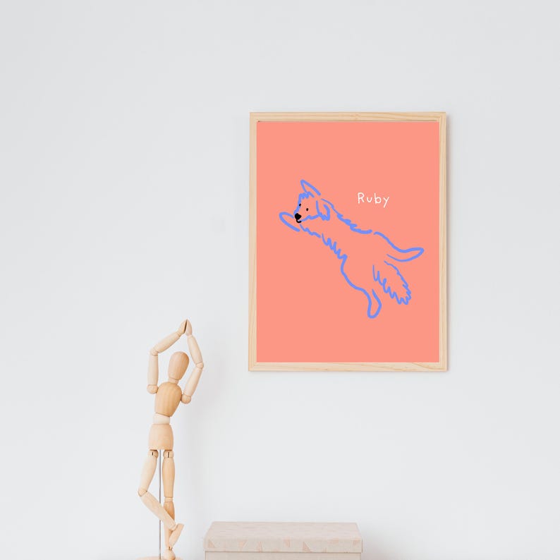 May include: A framed art print featuring a blue line drawing of a dog leaping across a coral background. The dog's name, "Ruby," is written in a small font above the dog. The frame is made of light-colored wood.