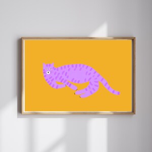 May include: A framed art print featuring a stylized illustration of a purple cat on a yellow background. The cat is depicted in a playful pose with a dotted pattern. The artwork is set within a wooden frame, adding a touch of warmth to the design.
